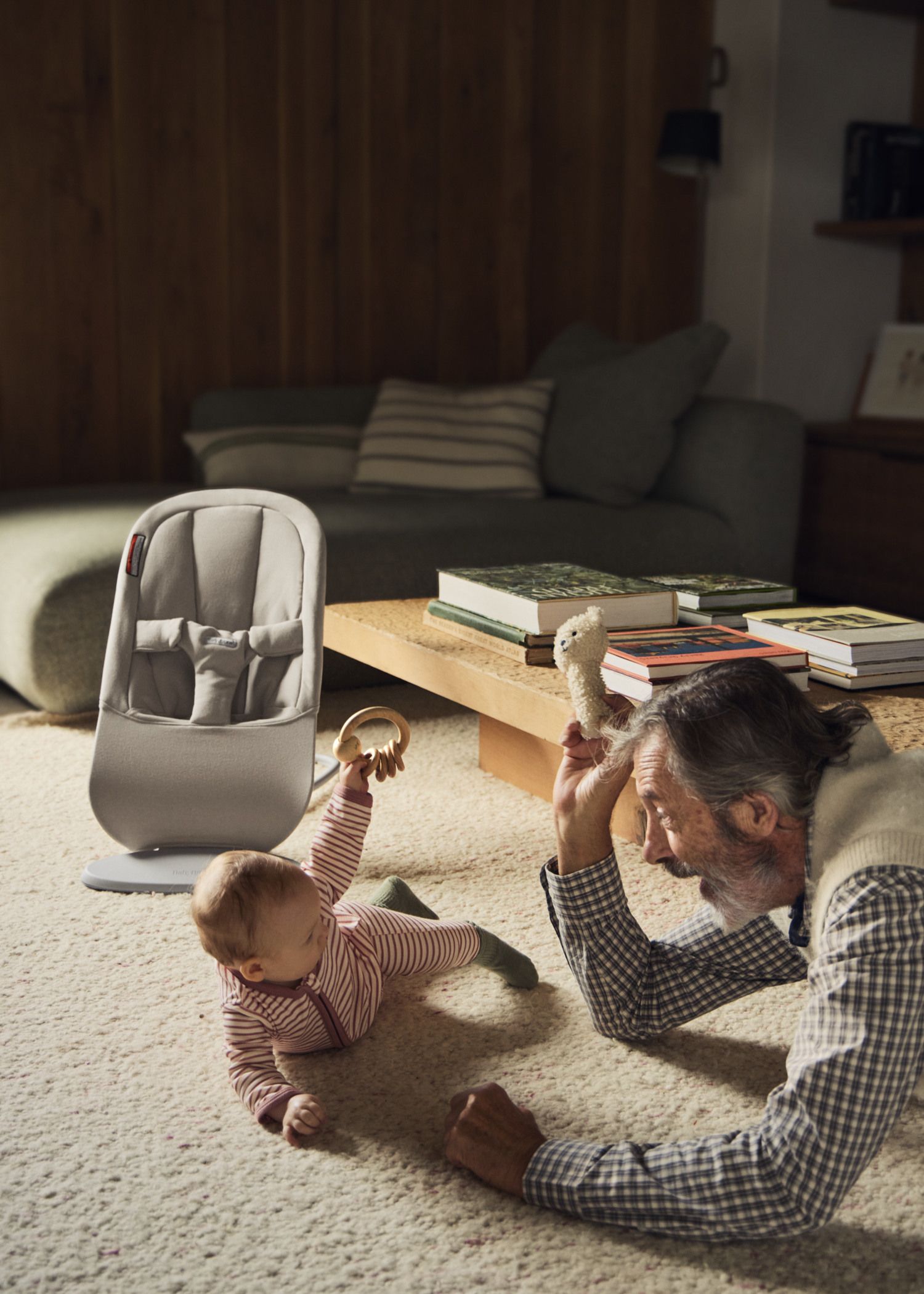 A baby in striped pajamas plays on the floor with a smiling elderly man who holds a stuffed toy. A grey baby chair and books are in the background.