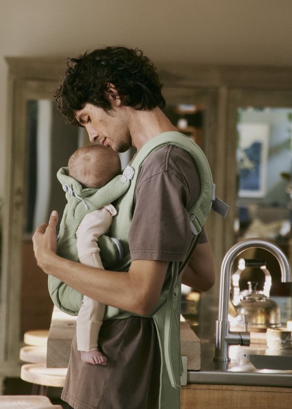 A person gently holding a baby in a green carrier in a cozy, warmly lit kitchen setting.
