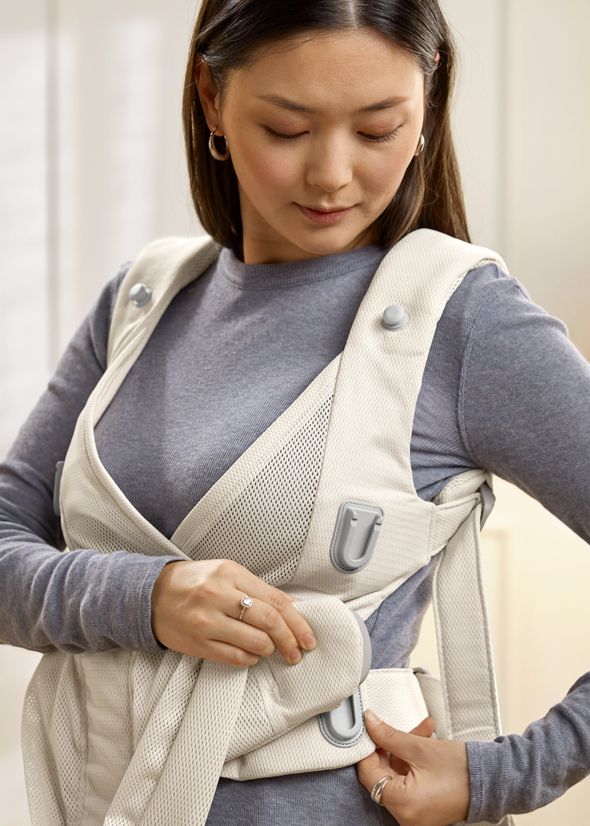 A woman wearing a gray top adjusts a beige baby carrier strapped around her, focusing on securing a buckle.