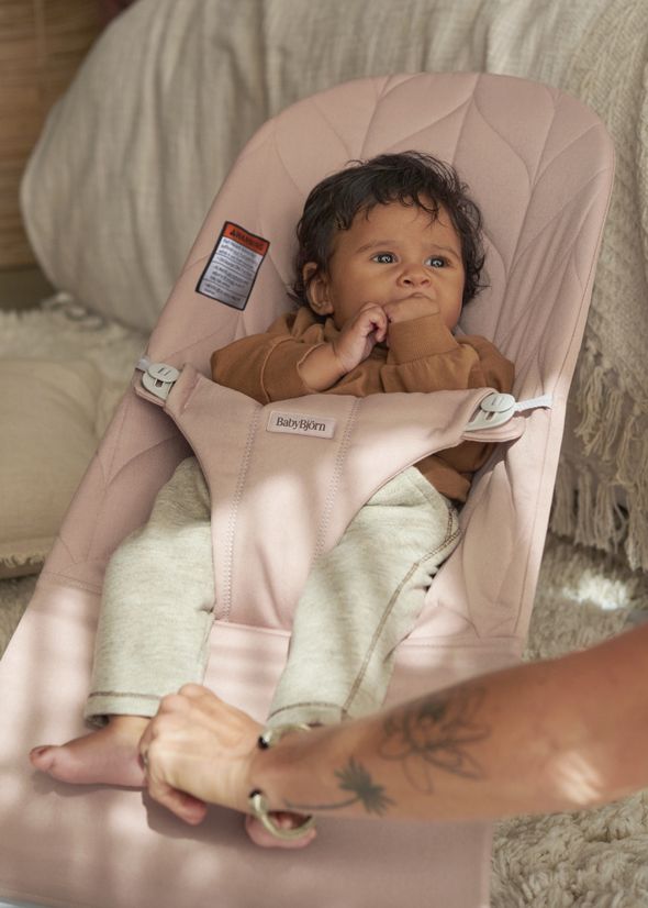 Baby in a pink bouncer wearing a brown shirt and beige pants, with an adult's hand gently holding the bouncer's edge.