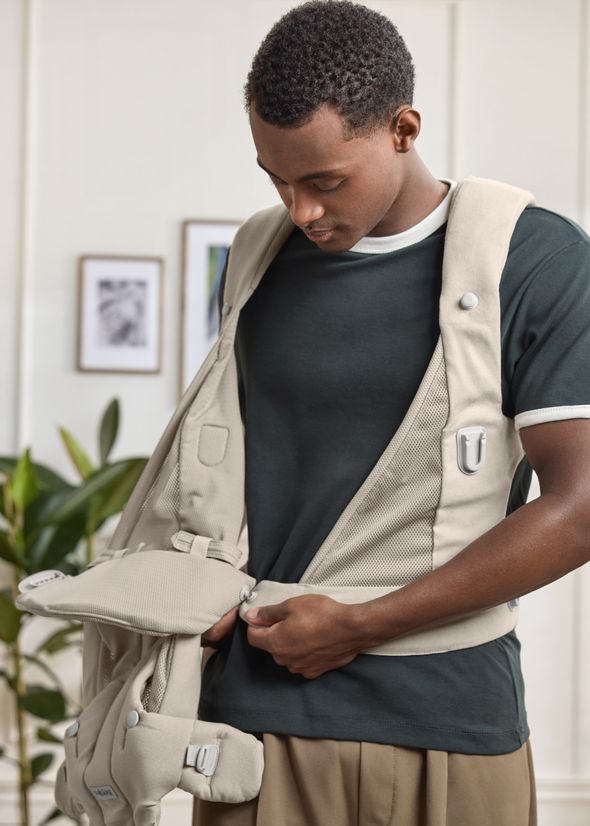A person wearing a dark T-shirt adjusts a beige baby carrier in a light room with framed pictures and plants in the background.
