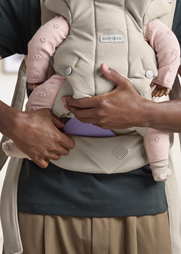 A person wearing a baby carrier with a baby in pink pajamas. The person's hands are adjusting the carrier strap.