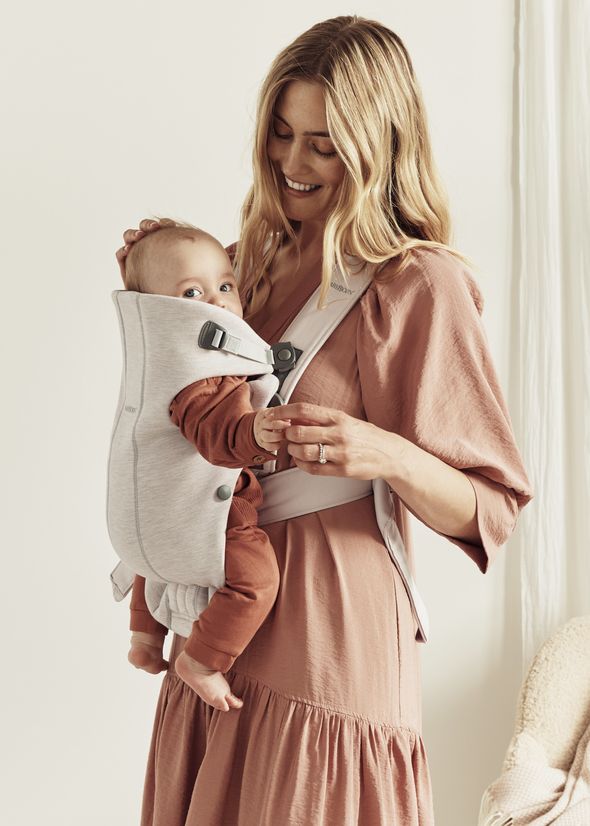 A woman smiling at a baby in a front-facing carrier. The baby is looking forward, and the woman is dressed in a soft pink dress.
