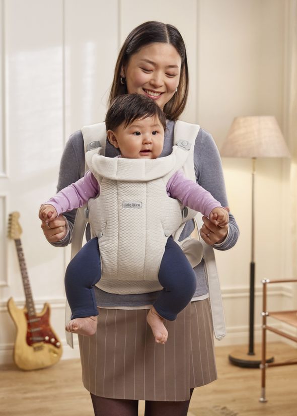 A woman smiling at a baby in a baby carrier. The baby is wearing a pink top and blue pants. A guitar and lamp are in the background.
