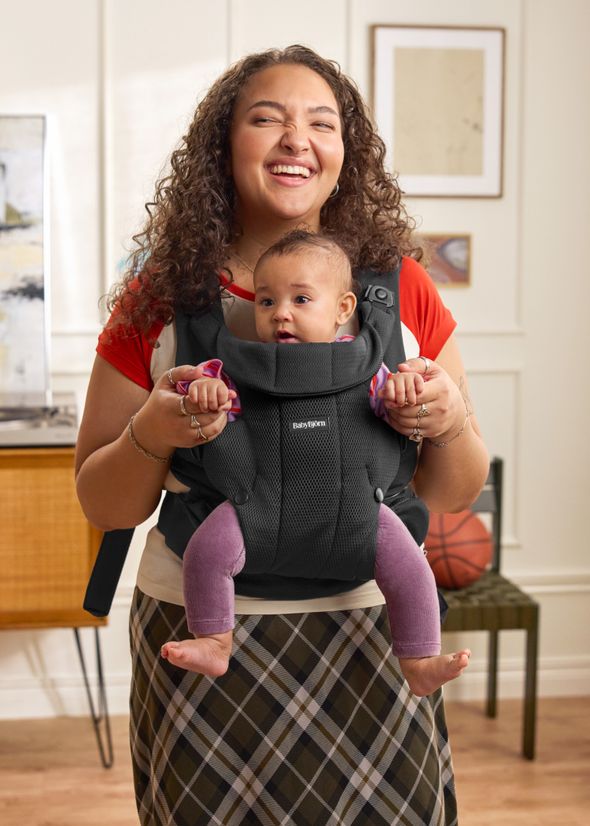 A woman smiling and holding a baby in a baby carrier. The baby is looking forward, also smiling. They are indoors in a warmly decorated room.