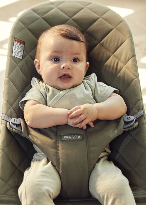 A baby in a green onesie sits in a quilted, olive-green baby chair, clasping hands, with sunlight softly illuminating the scene.