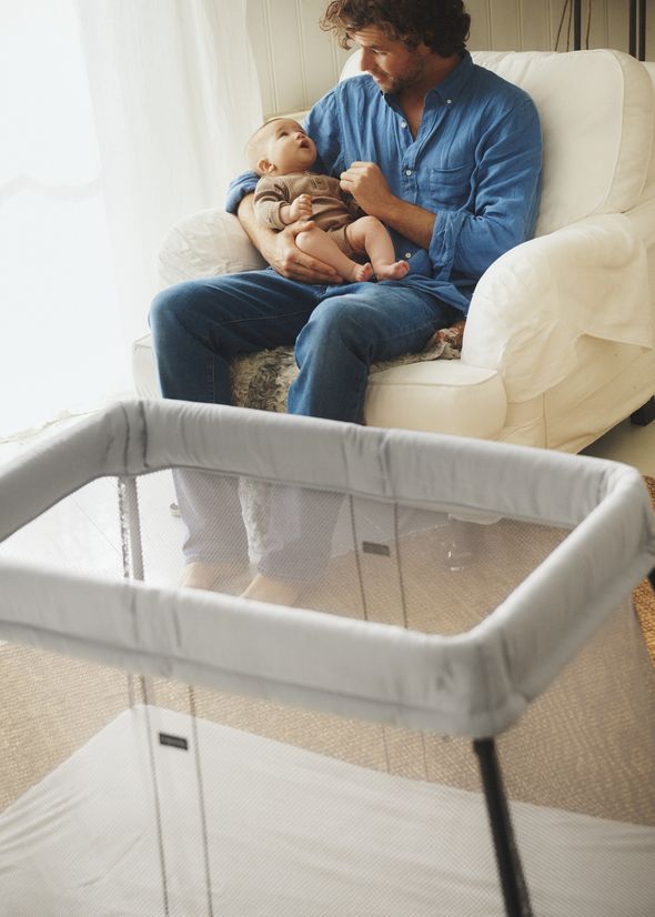 A man in a blue shirt sits on a white chair, holding a baby in his lap, with a travel crib in the foreground.