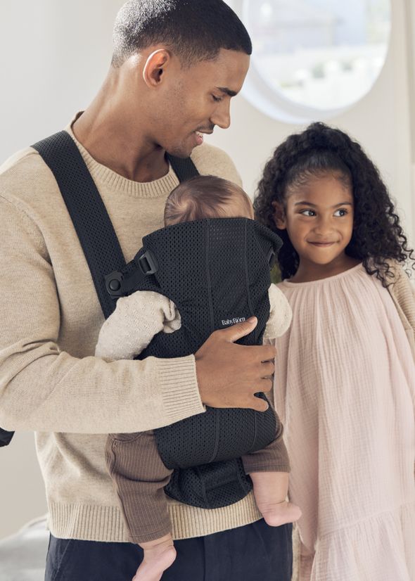 Man wearing a beige sweater smiles at baby in a black carrier, standing next to a young girl in a pink dress, inside a bright room.