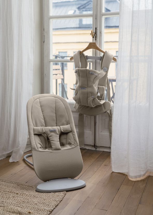 A beige baby bouncer on the floor and a matching baby carrier hanging on a chair in front of a window with sheer curtains.
