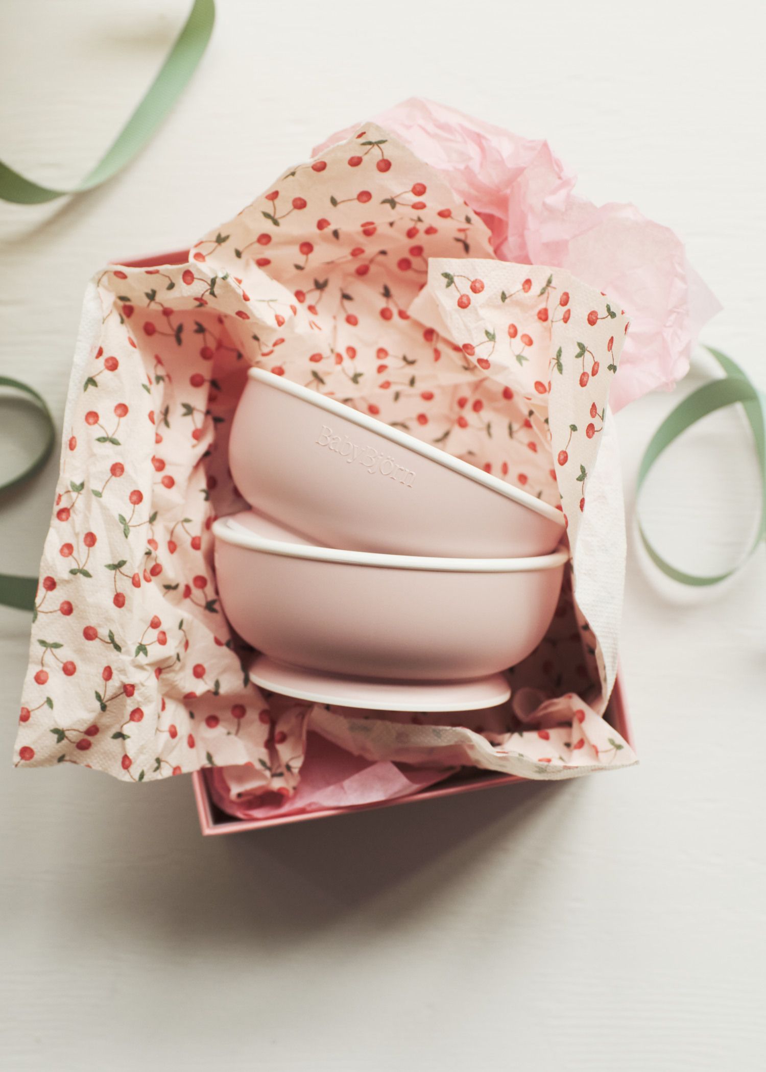 Two pink bowls stacked in a gift box with cherry-patterned tissue paper and green ribbons on a light background.