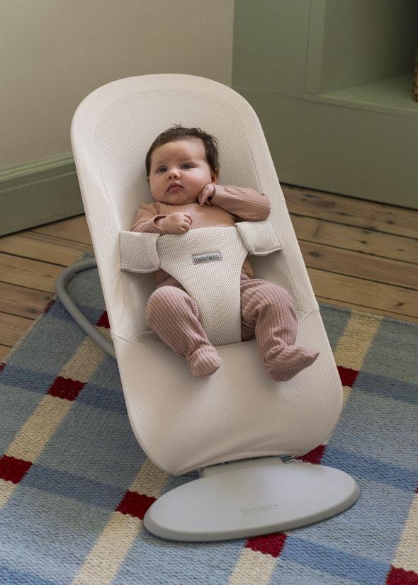Baby in a pink outfit seated in a white baby rocker on a blue and red checkered rug, looking up.