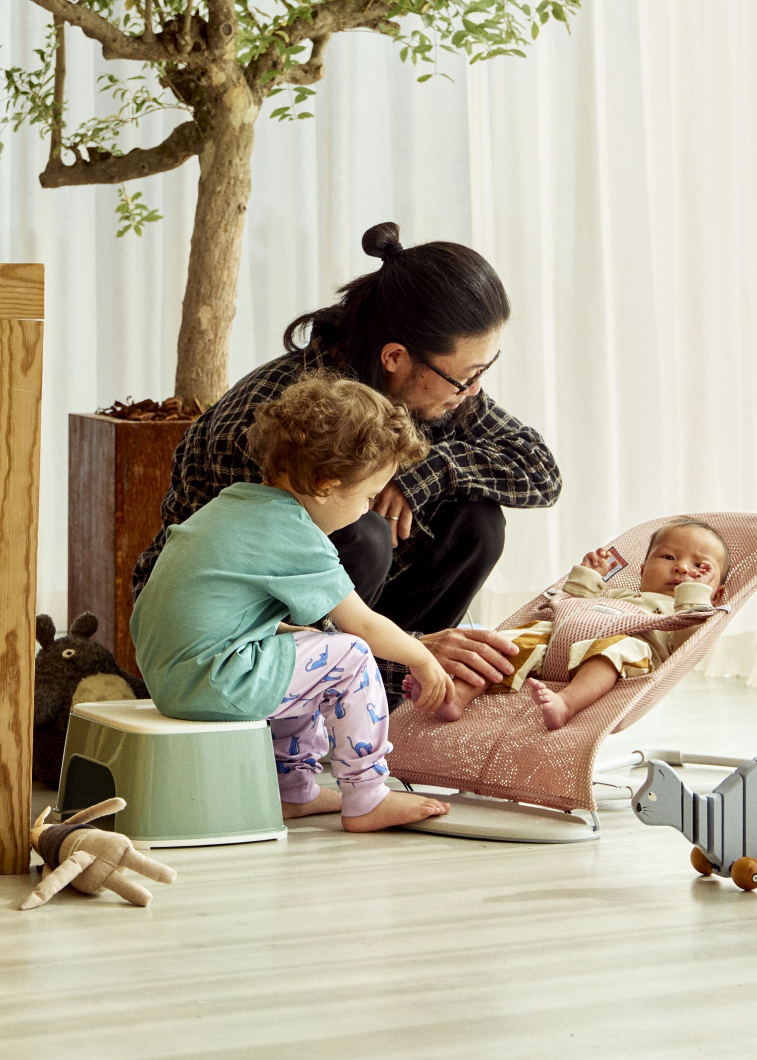 A man and a child sit on the floor, looking at a baby in a bouncer. A toy and a plant are nearby.
