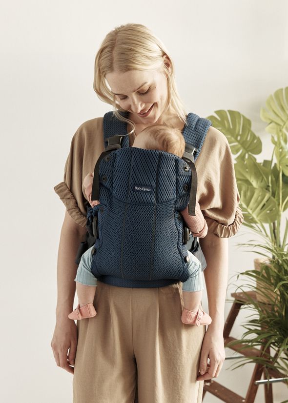 A woman smiling at a baby in a blue carrier. The baby faces her chest. Both are indoors, with a green plant in the background.
