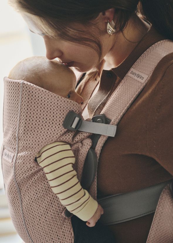 A woman gently kisses a baby's head while carrying them in a pink baby carrier. The baby wears a striped outfit.