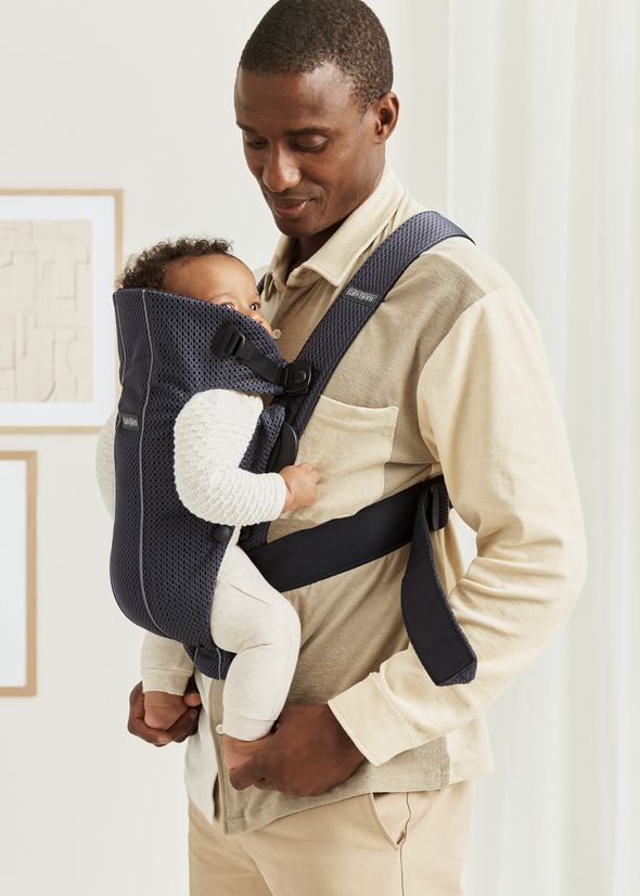 A man carrying a baby in a blue carrier, the baby is wearing a beige outfit, and they are in a softly lit indoor setting.