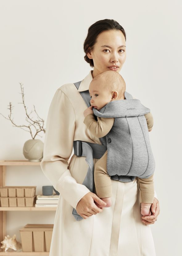 A woman wearing a white blouse holds a baby in a gray carrier. They stand indoors, with a minimalist shelf and decor in the background.