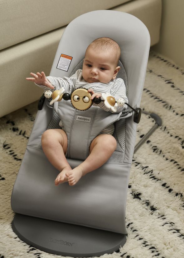 Baby in a grey bouncer reaching out to a toy bar, sitting on a patterned rug.