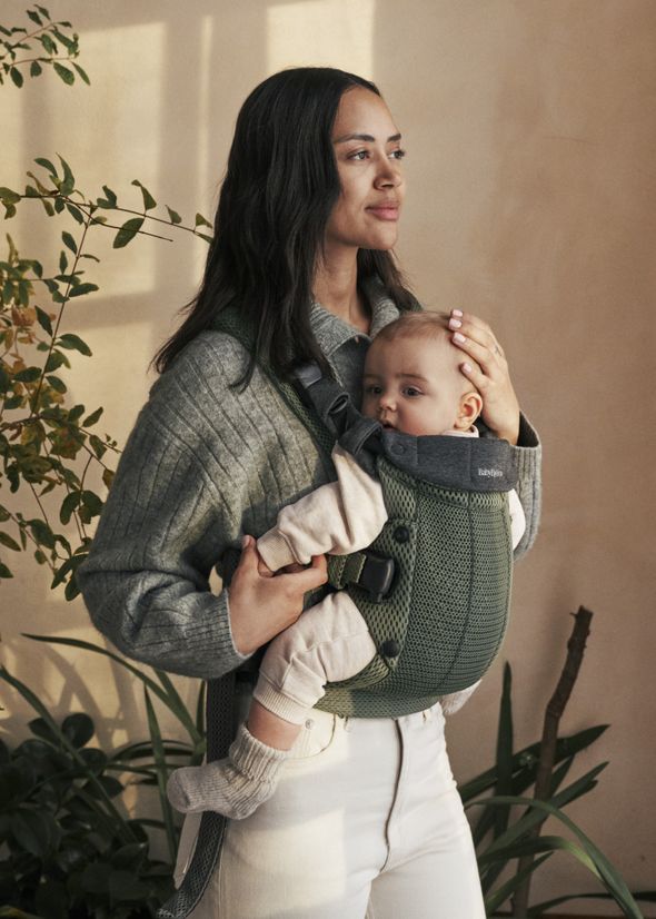 A woman wearing a gray sweater carries a baby in a green baby carrier, standing indoors near plants.