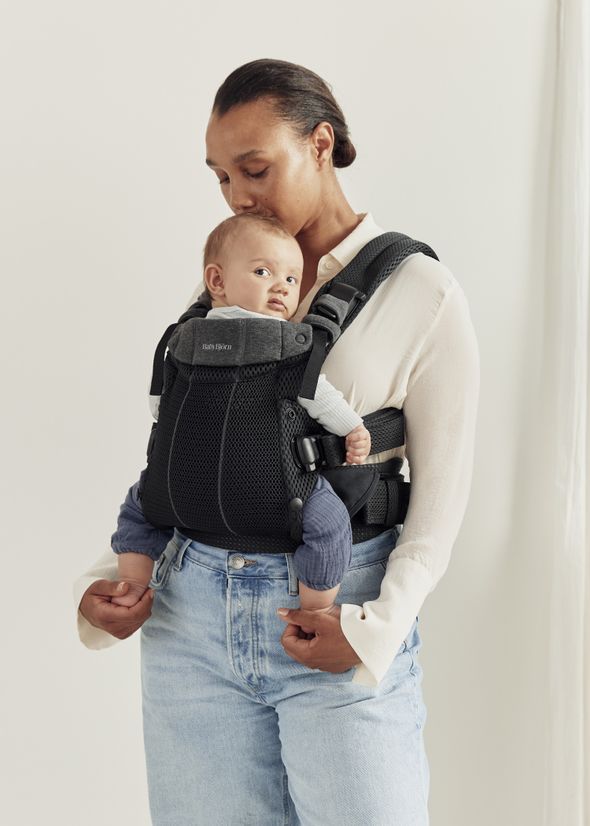 A person wearing a white shirt holds a baby in a gray and black baby carrier, gently kissing the baby's head.