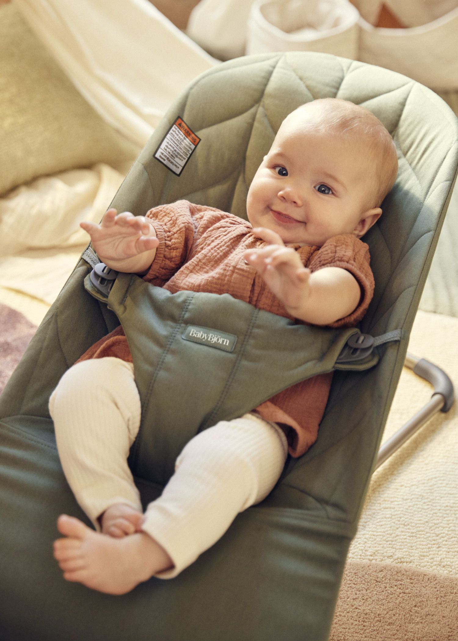 Smiling baby in a brown shirt and white pants, sitting in a green baby bouncer, reaching out with both hands.