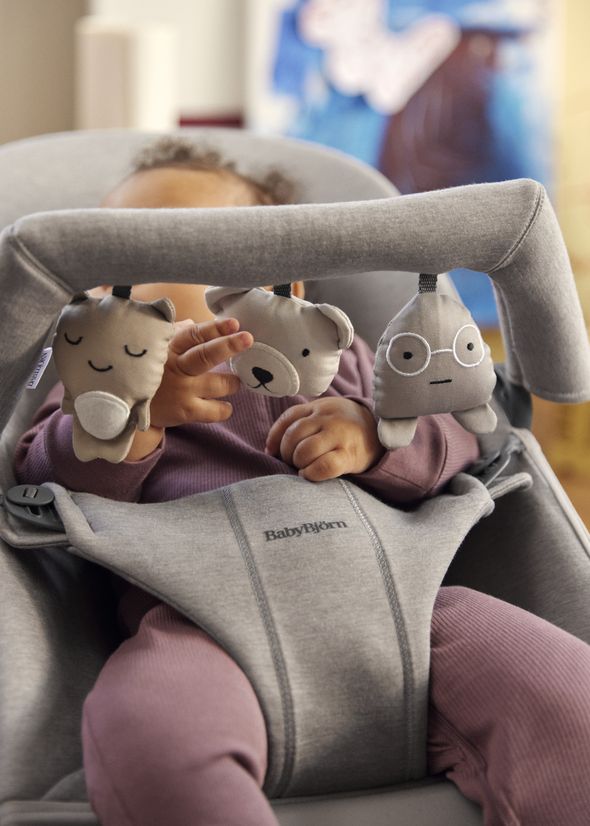 Baby in a gray bouncer wearing a purple outfit, reaching for plush toys attached to the bouncer's handle.