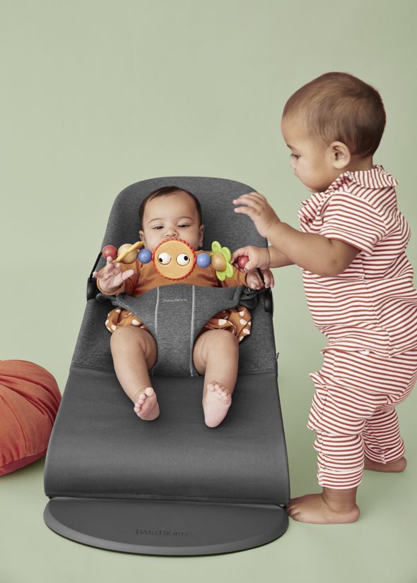 Two babies, one sitting in a baby bouncer with a toy, the other standing beside, wearing striped pajamas, on a light green background.