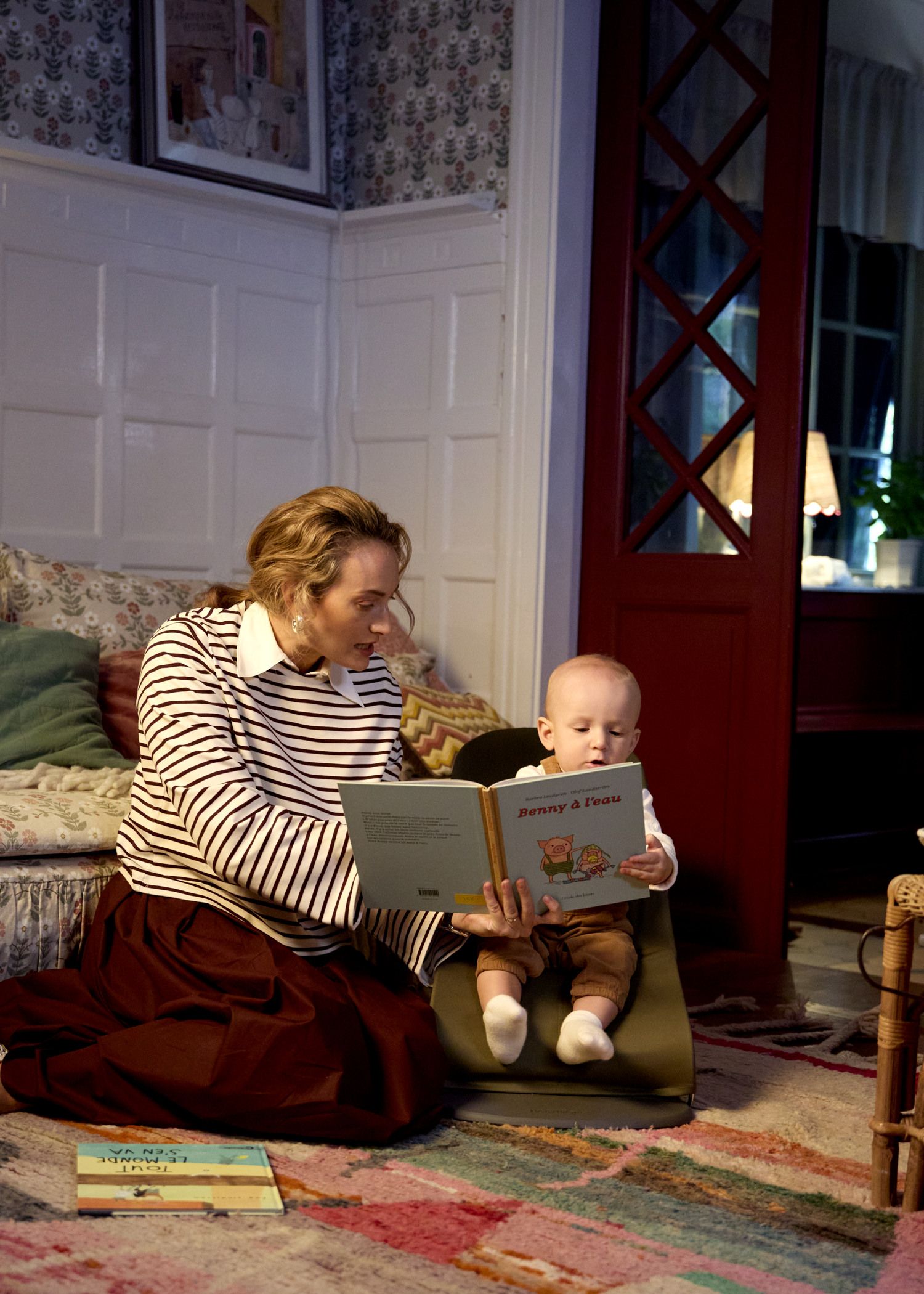 A woman and a baby are sitting on the floor in a cozy room, reading a book together. The baby is seated on a cushion, holding the book.