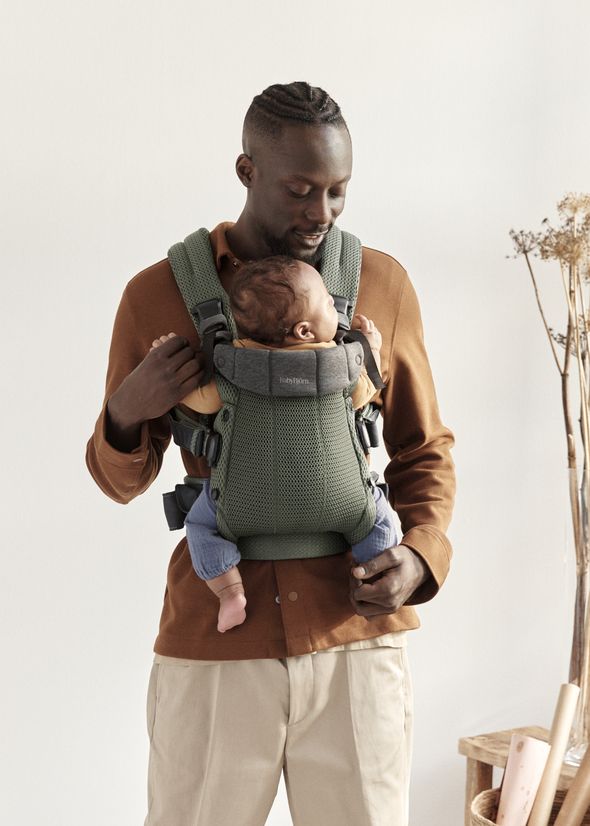 A man stands holding a baby in a green carrier. He wears a brown shirt and beige pants, looking at the baby affectionately.