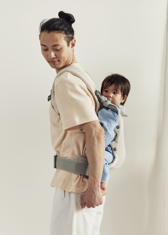A person is wearing a light-colored outfit, carrying a baby in a blue outfit in a gray baby carrier on their back, against a plain background.