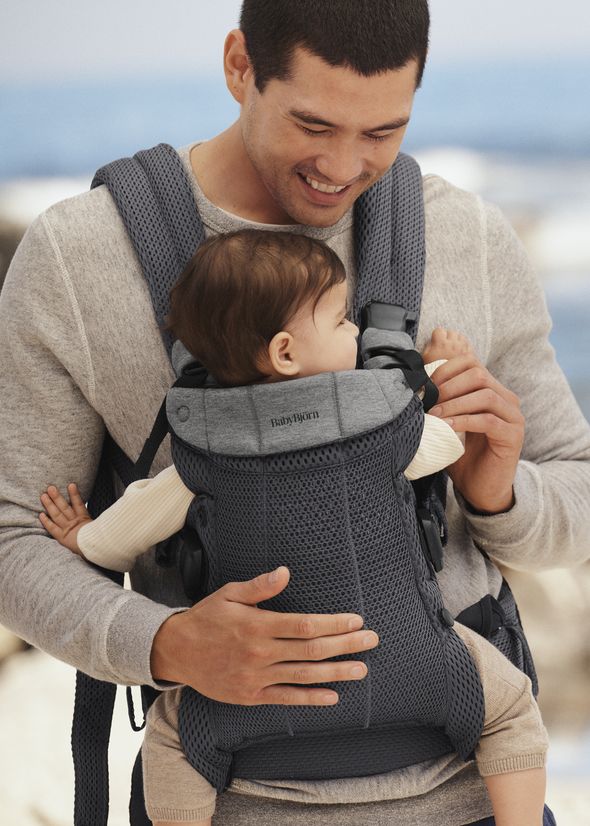 A smiling man holds a baby in a gray baby carrier, both dressed in light-colored clothing, with a blurred outdoor background.