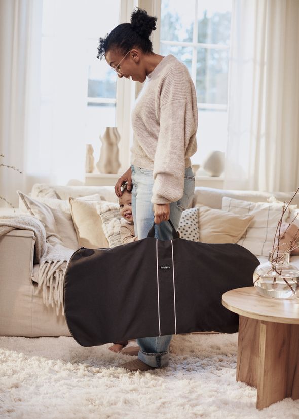 A woman in a cozy living room holds a large, black BabyBjörn bag while a child peeks from behind it, smiling.