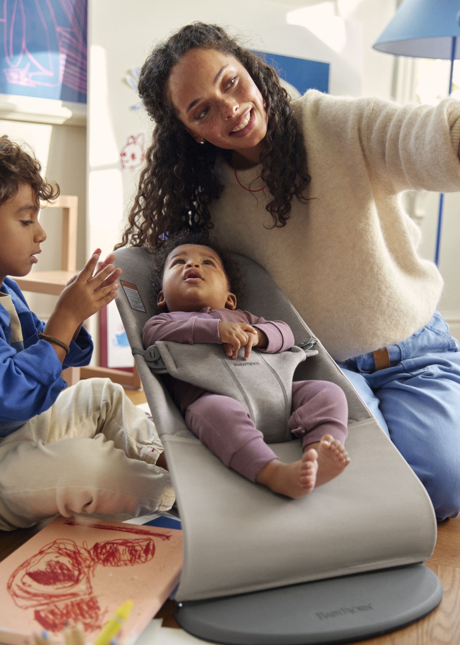 A woman smiles while sitting with two children; one child is in a baby bouncer, and the other is drawing with crayons.