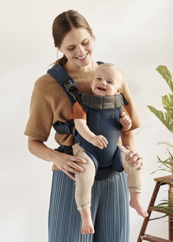 A woman smiles warmly at a baby sitting in a blue carrier she is wearing, against a neutral background with a plant visible on the side.