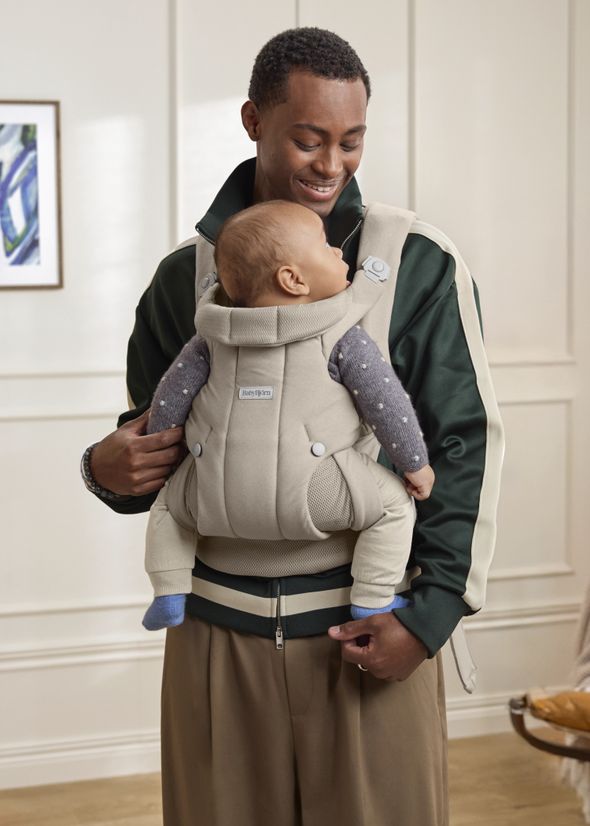 A smiling man is holding a baby in a beige baby carrier, standing indoors with a painting on the wall behind them.