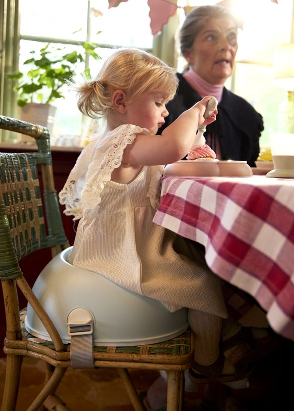A young child in a high chair eats at a table with a red checkered tablecloth, while an adult sits nearby.