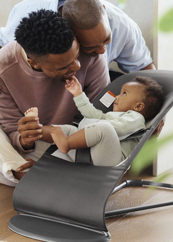 Two adults smile and interact with a baby sitting in a gray baby bouncer, creating a warm and joyful atmosphere.