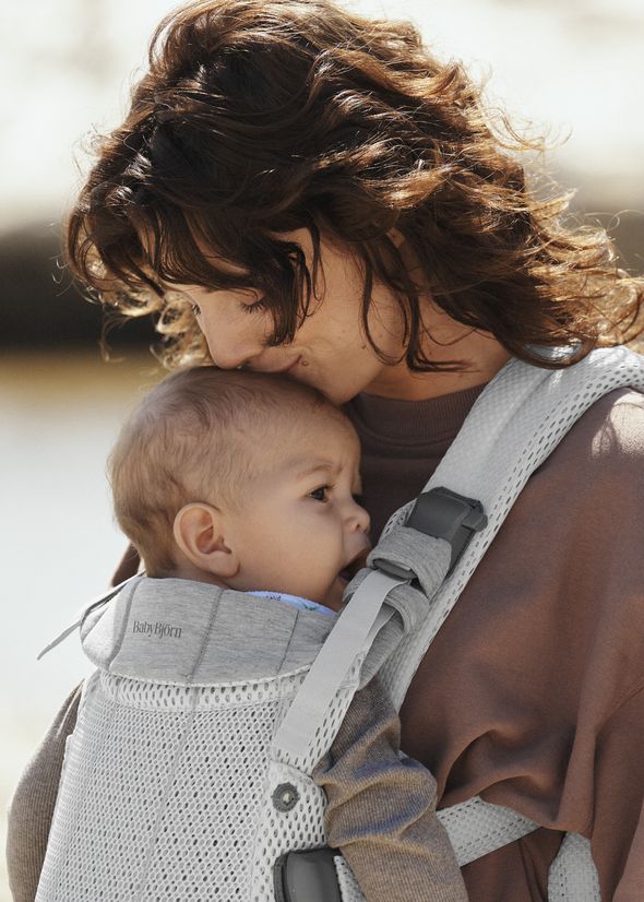 A woman gently kisses the head of a baby she is carrying in a gray BabyBjörn carrier, both appearing content and relaxed.