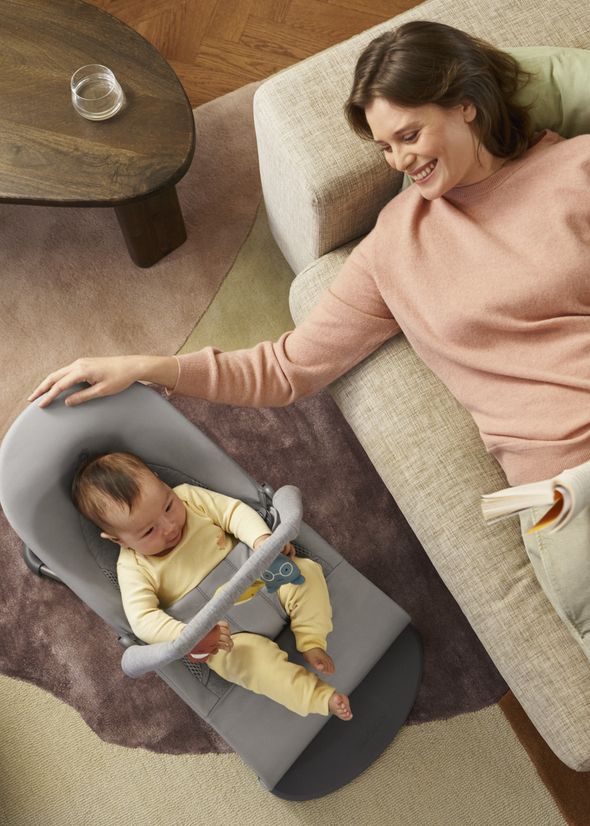 Woman relaxing on a couch, smiling at a baby in a gray bouncer on the floor; a cozy living room scene.