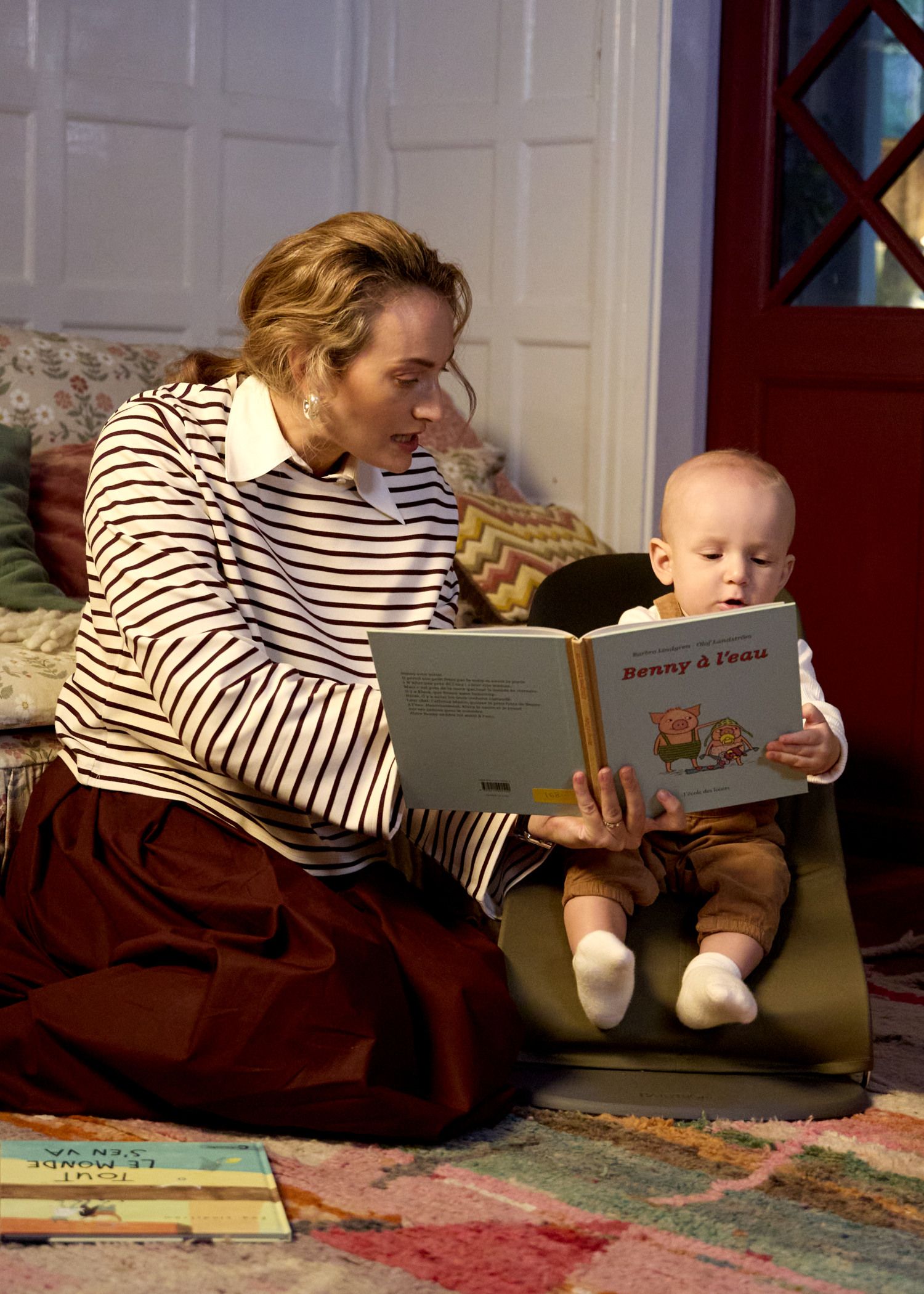 A woman and a baby sit on the floor reading a book together in a cozy room with colorful decor.