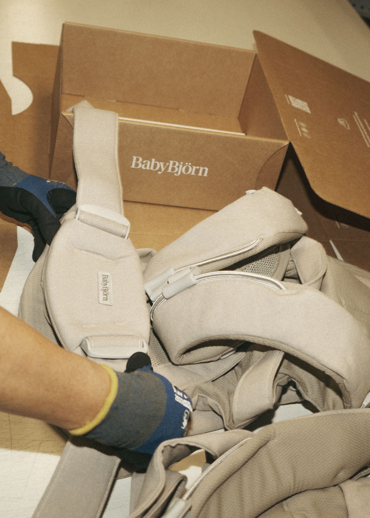 A person wearing gloves assembling a beige BabyBjörn baby carrier beside an open cardboard box.