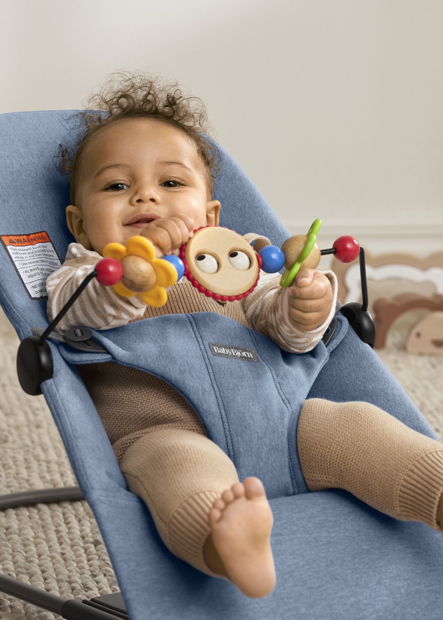 A baby smiling and playing with colorful toys in a blue BabyBjörn bouncer.