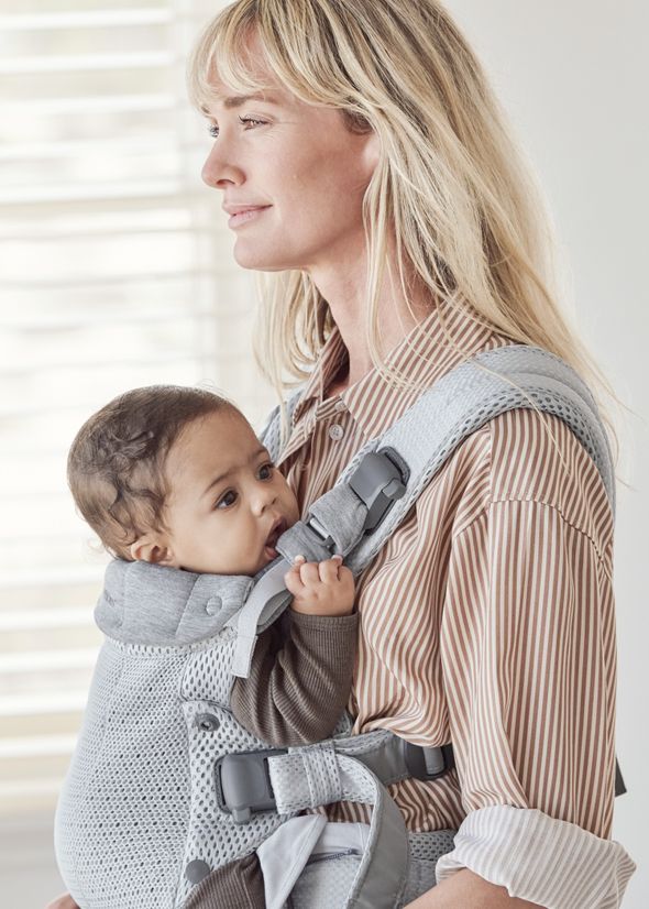 A woman with long blonde hair wearing a striped shirt carries a baby in a gray baby carrier. The baby looks to the side, gripping the carrier.