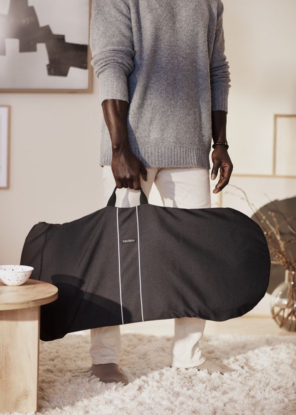 A person in a gray sweater and white pants holds a black transport bag for bouncer with a handle, standing in a cozy, art-decorated room with a light rug.