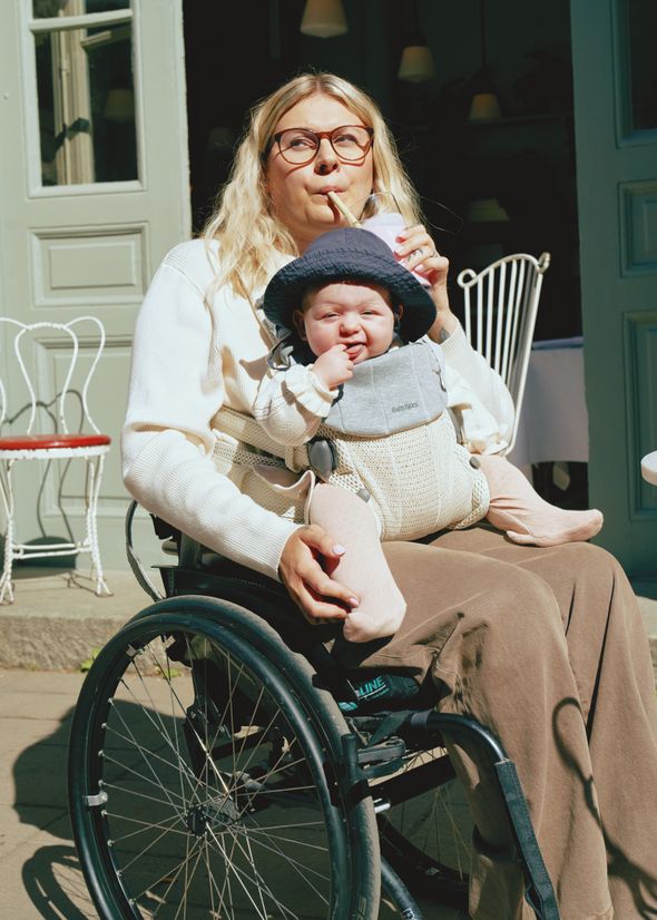 Woman in a wheelchair sipping a drink holds a baby in a carrier. The baby wears a hat and looks content. They are outside a café.