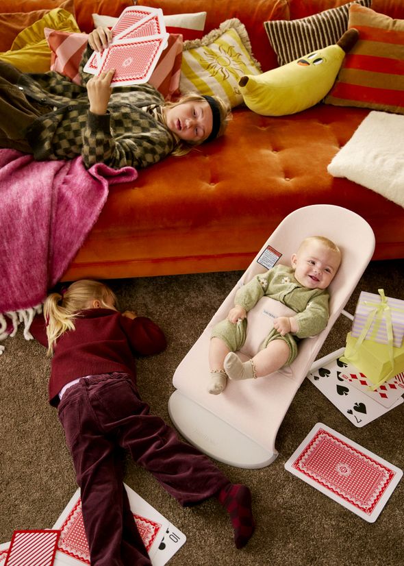 A baby in a bouncer and a child lie on the carpet, surrounded by oversized playing cards, with an adult relaxing on an orange sofa.
