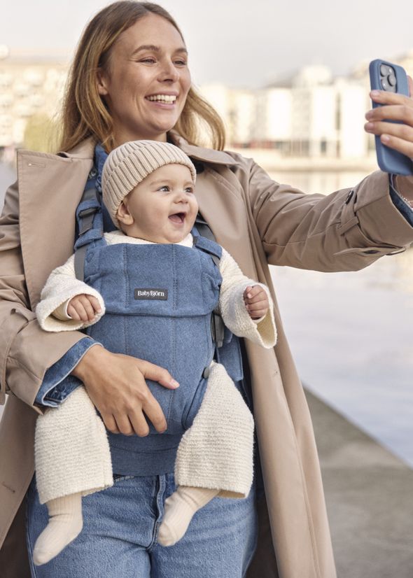 Smiling woman taking a selfie outdoors, holding a cheerful baby in a blue carrier. Both are dressed warmly, with a cityscape in the background.