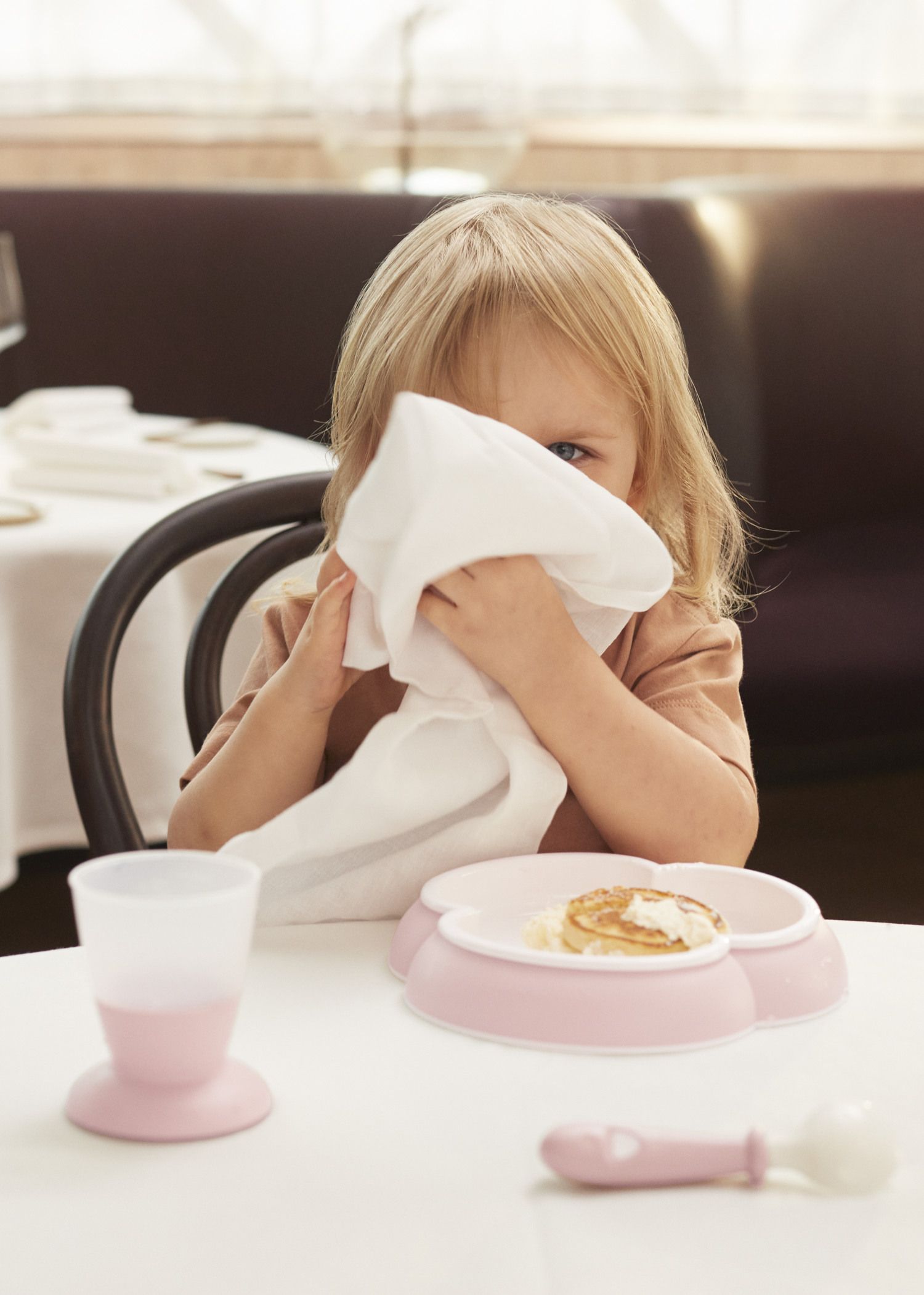 A child in a brown shirt playfully hides behind a napkin at a table set with a pink plate and cup.