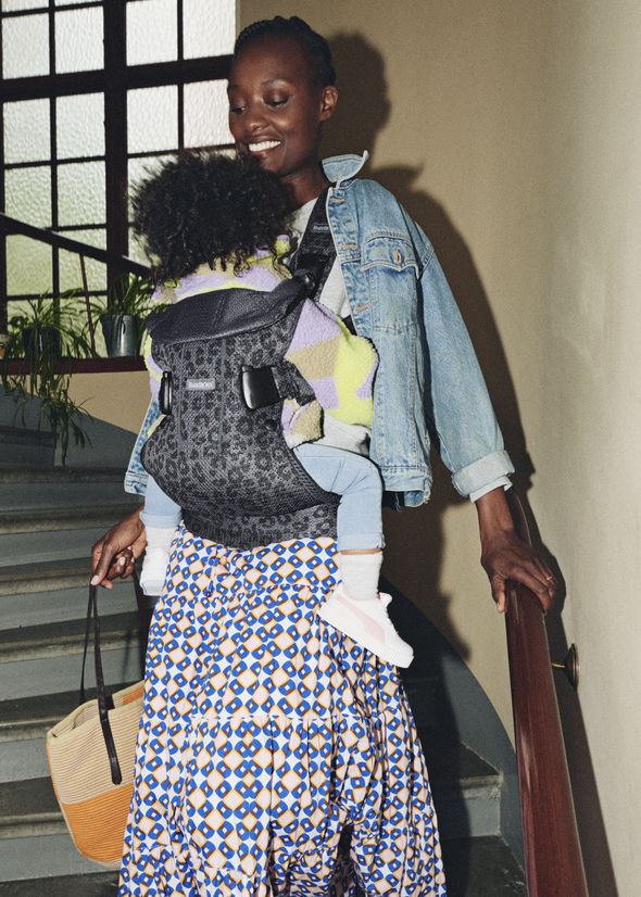 Smiling woman in denim jacket carries a baby in a carrier on her chest, walking down stairs with a woven bag.