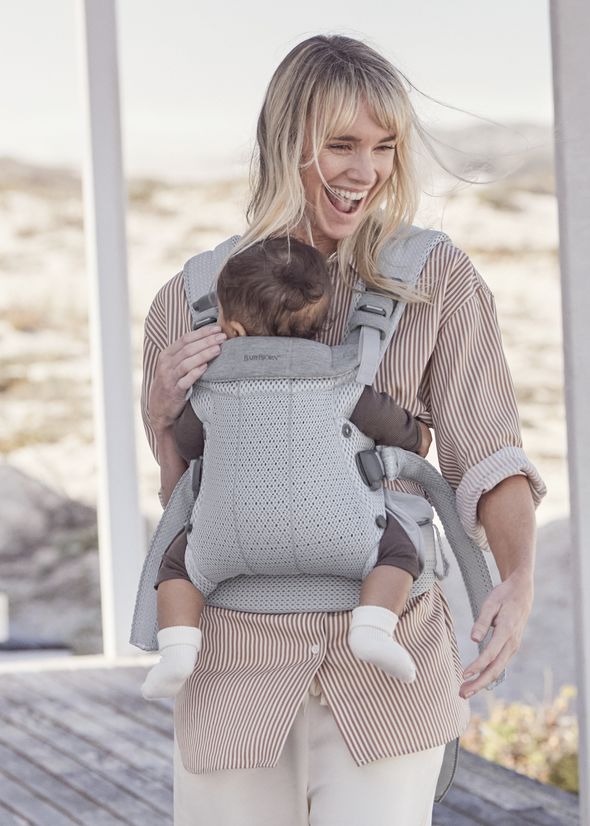 Smiling woman outdoors carrying a baby in a gray baby carrier. She wears a striped shirt and white pants.