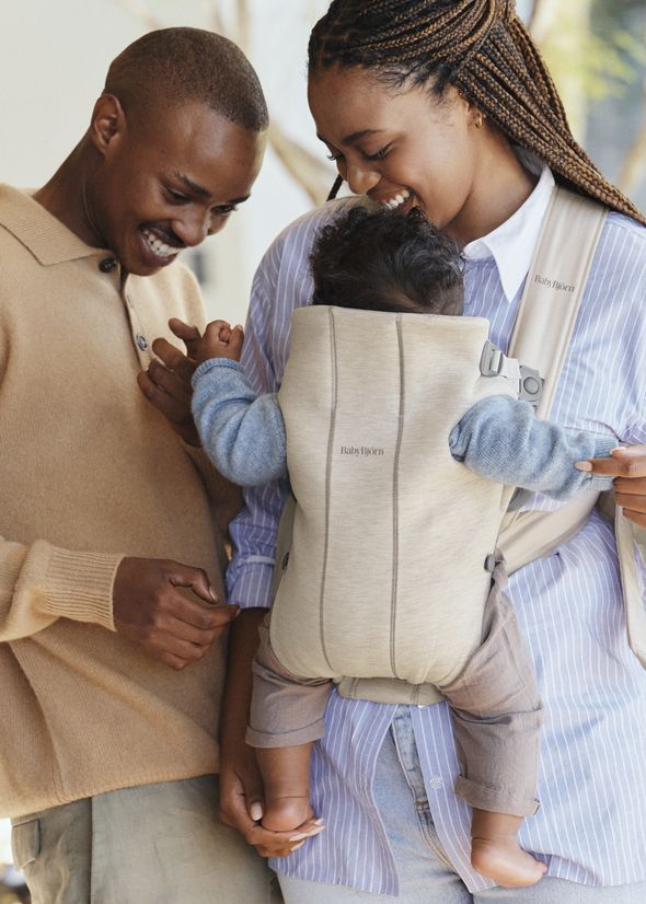 A smiling couple happily looks at their baby in a beige carrier. The father holds the baby's hand while the mother supports the carrier.
